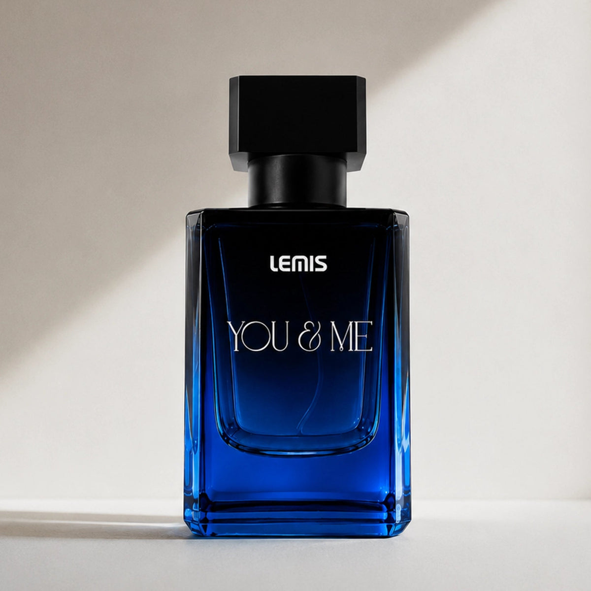 A rectangular blue glass perfume bottle with a black cap, labeled 'LENIS YOU & ME', standing on a light surface against a warm beige wall with soft diagonal shadows.