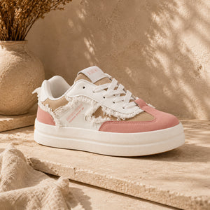 A low-top sneaker with a white and dusty pink color scheme, featuring distressed raw-edge detailing on the side, white laces, and a chunky white sole, displayed on a textured stone surface.