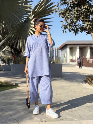 A woman wearing a light blue linen two-piece tunic and trouser set walks outdoors on a paved path. The set features a short-sleeve, V-neck tunic top and matching wide-leg pants.