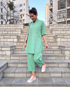 A woman wearing a light green two-piece linen set consisting of a short-sleeve tunic and wide-leg pants, walking down outdoor stone steps.