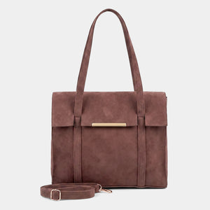 A structured brown suede tote bag with a front flap, a horizontal gold-tone metal bar accent, and a matching detachable shoulder strap, displayed against a light gray background.