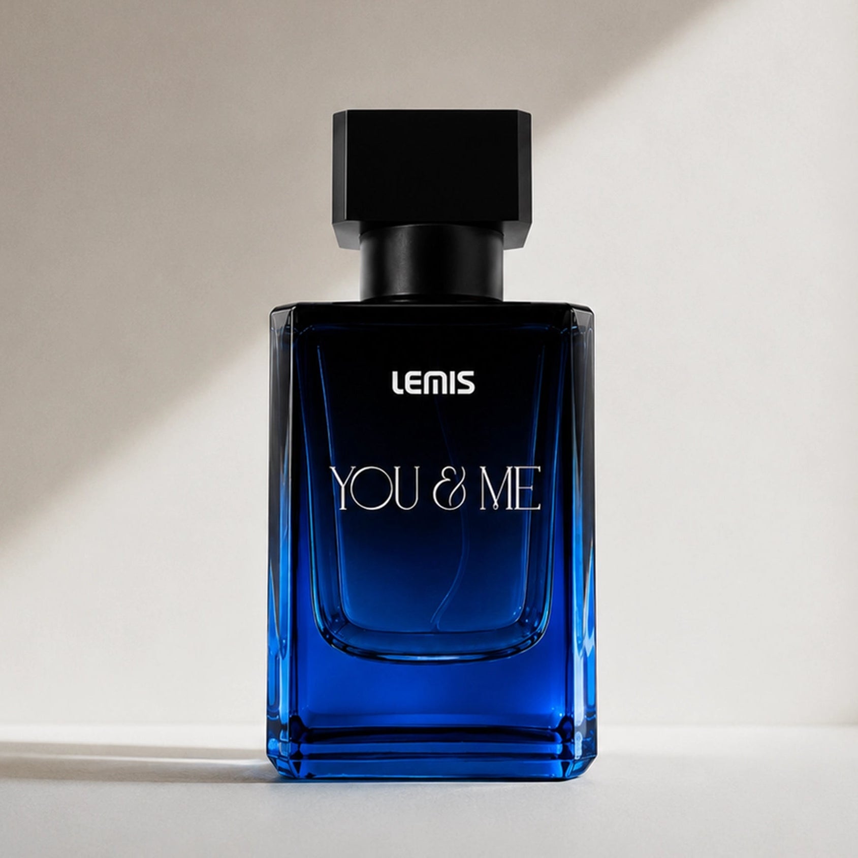 A rectangular blue glass perfume bottle with a black cap, labeled 'LENIS YOU & ME', standing on a light surface against a warm beige wall with soft diagonal shadows.