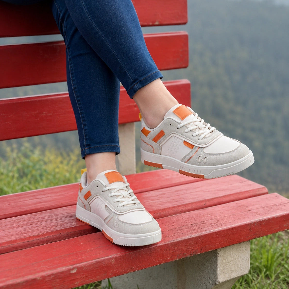A person sitting on a red wooden park bench wearing light gray and white low-top sneakers with orange accents, set against a blurred green mountain background.