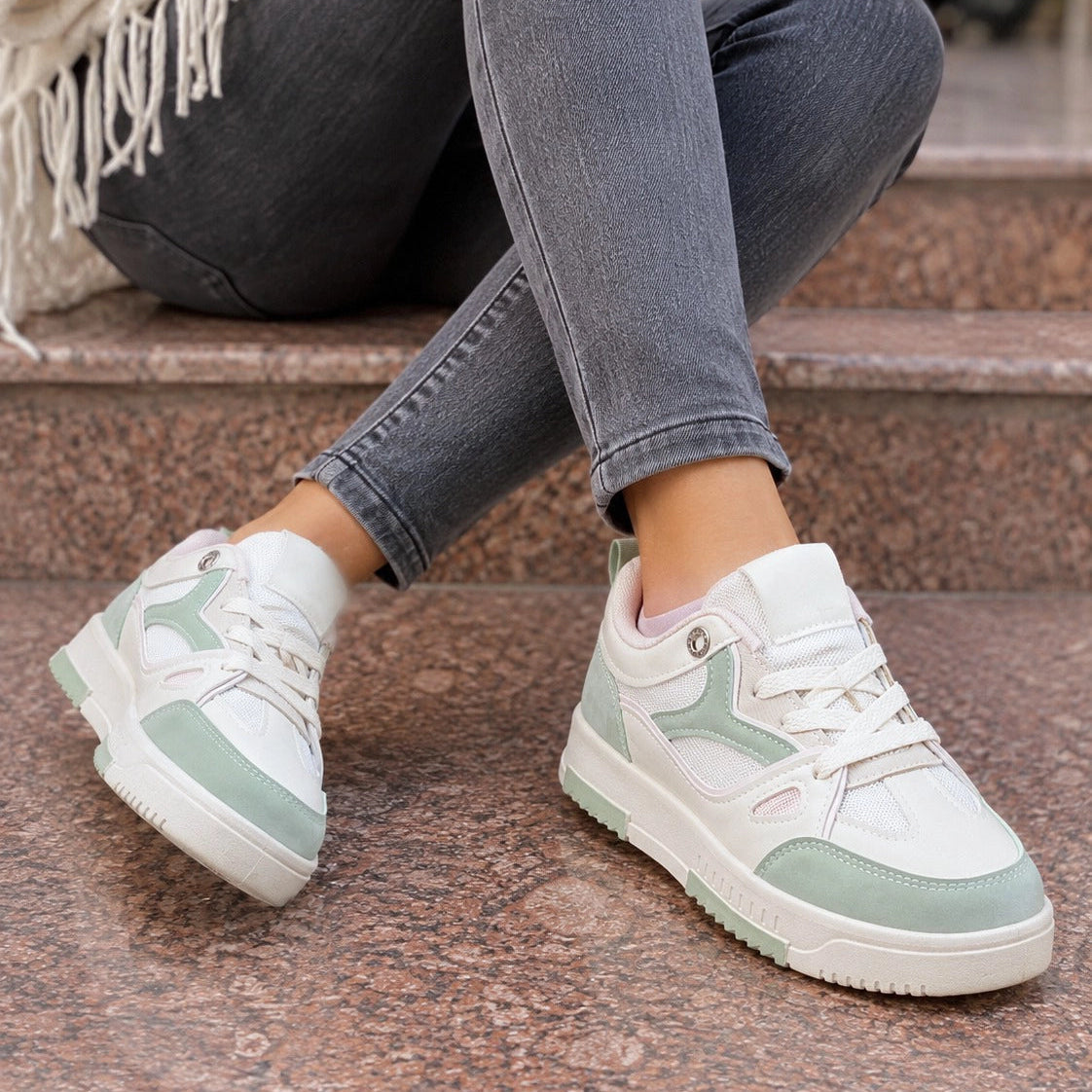White low-top sneakers with mint green accents worn by a person sitting on stone steps, paired with gray jeans.