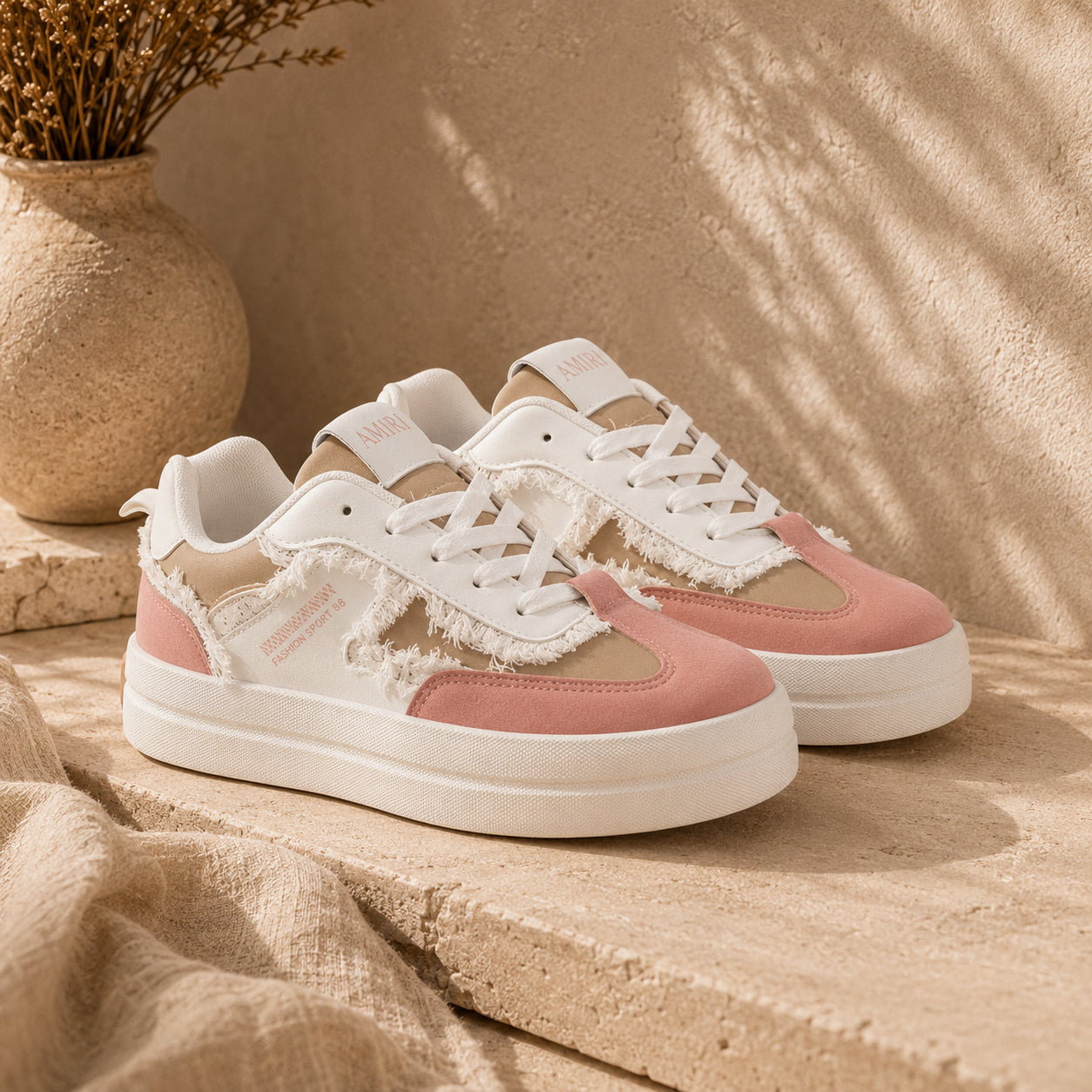 A pair of low-top platform sneakers in a dusty pink and white colorway, styled on a beige stone ledge with a textured wall and a vase in the background.