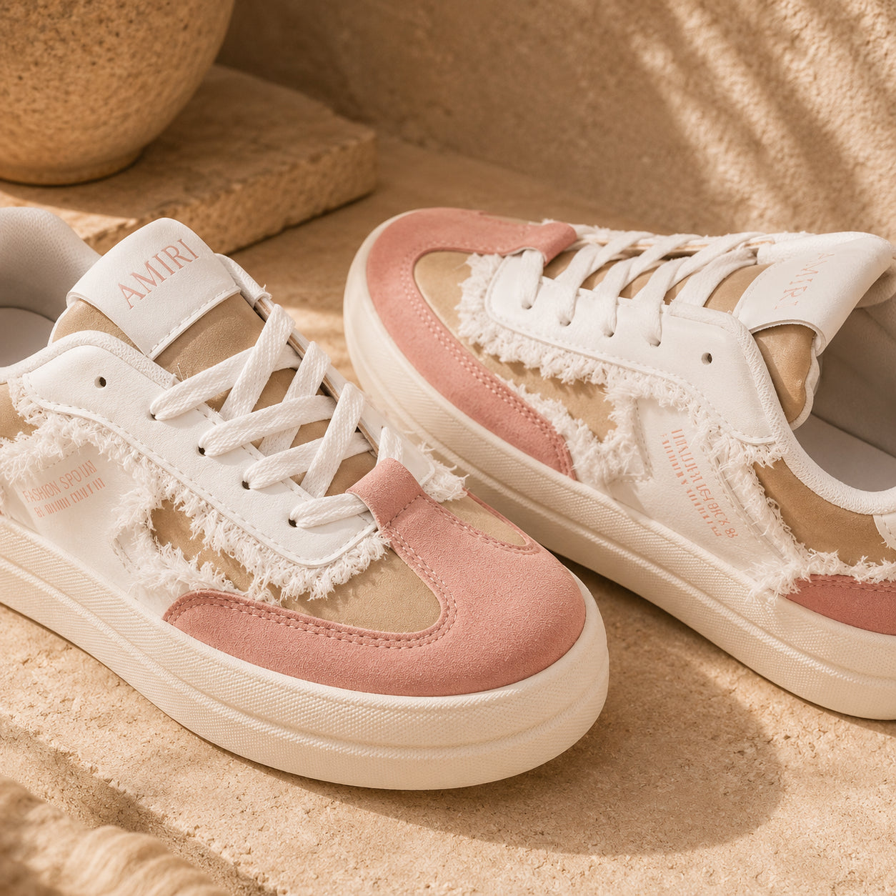 A pair of low-top sneakers in white and dusty pink suede with distressed cutout details, shown on a sandy beige surface with a warm, sunlit outdoor feel.