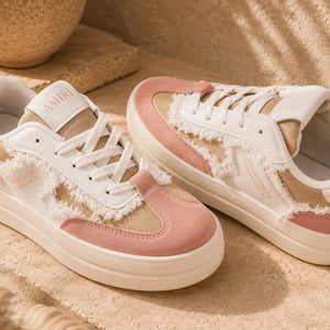 A pair of low-top sneakers in white and dusty pink suede with distressed cutout details, shown on a sandy beige surface with a warm, sunlit outdoor feel.