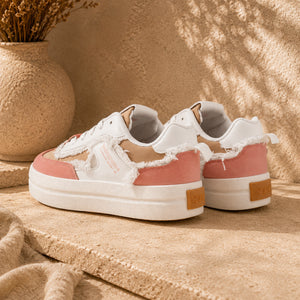 A pair of distressed platform sneakers in white and dusty pink, styled on a textured beige stone surface with a dried flower vase in the background.