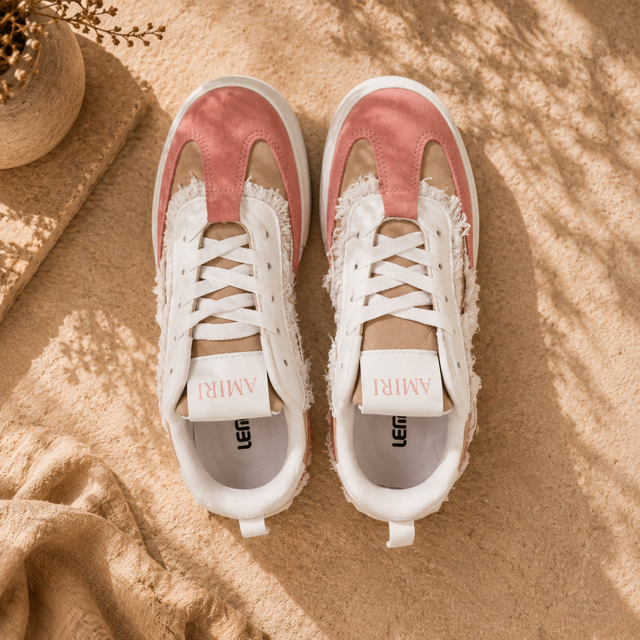 A pair of low-top sneakers shown from above on a sandy surface with soft shadows. The shoes are white with dusty pink suede panels and distressed frayed detailing around the sides and laces.