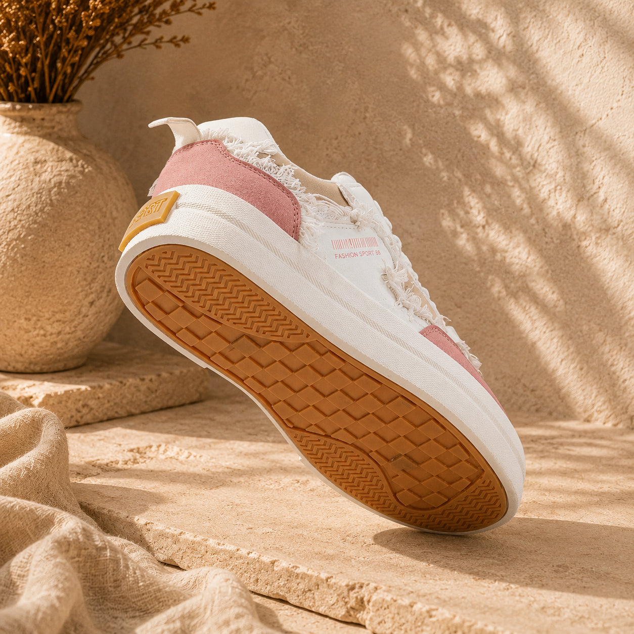 A single low-top sneaker shown at an angle, displaying its white sole, tan gum outsole, and pink and white lace-up upper. The shoe is set against a warm, textured beige background with a rustic ceramic vase and soft shadows.