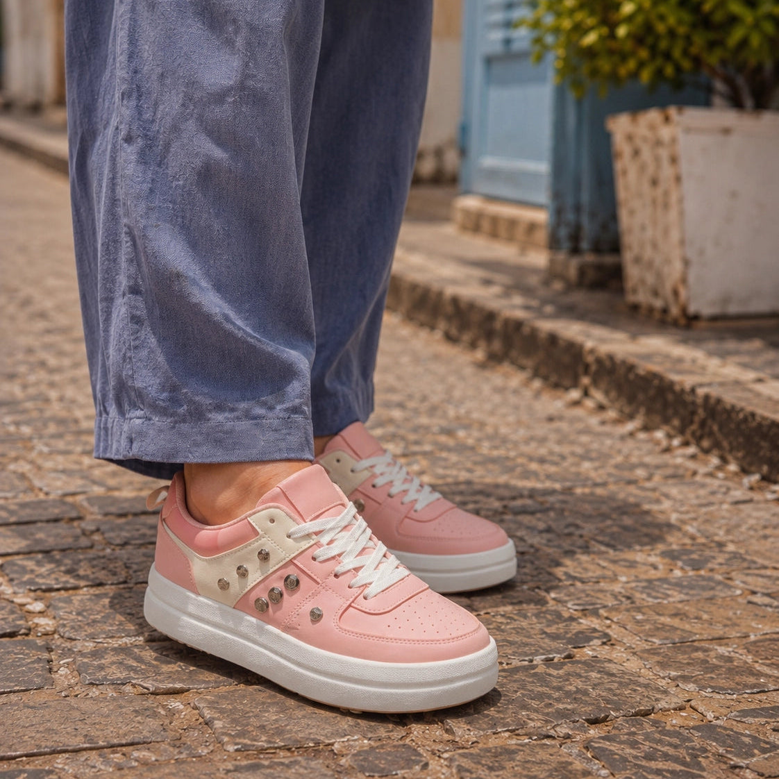 A person wearing pink low-top sneakers with white soles and decorative silver studs, paired with loose blue pants, standing on a cobblestone street.