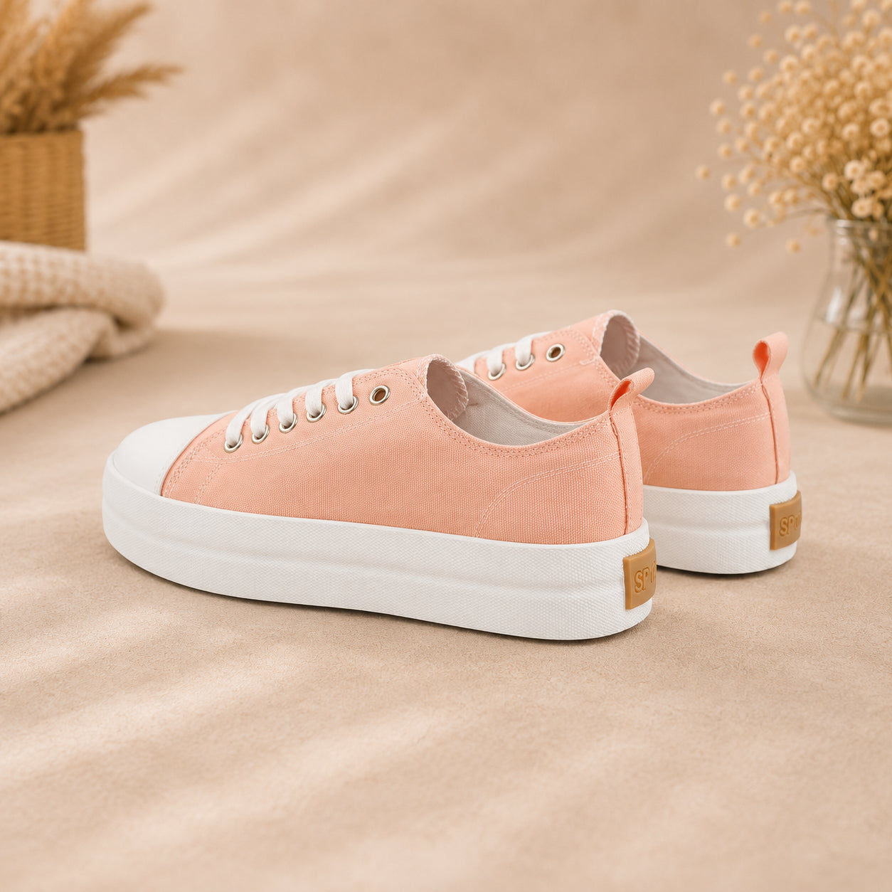 A pair of low-top canvas sneakers in a soft peach-pink color with white laces, white rubber soles, and small tan heel tabs, displayed on a beige fabric surface with dried floral props in the background.