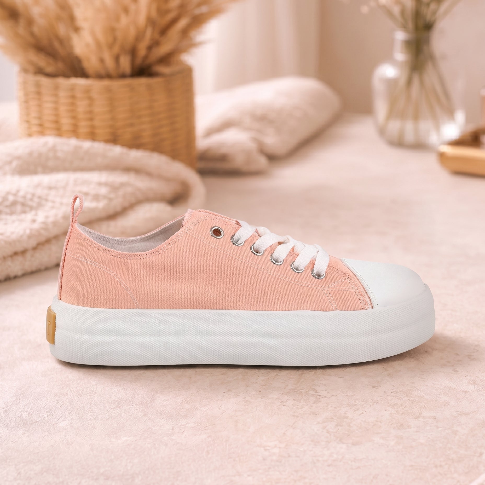 A low-top canvas sneaker in a soft peach-pink color with a thick white sole and white laces, styled on a beige textured surface with a cozy background of dried grasses and a glass vase.