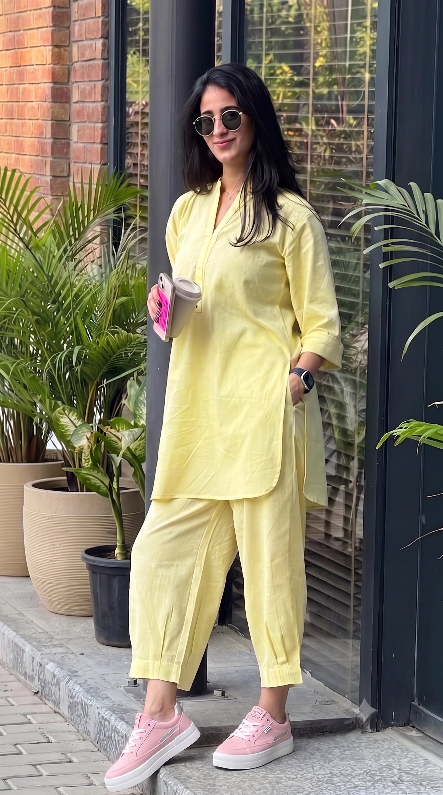 A woman wearing a light yellow two-piece kurta set with matching pants, sunglasses, and pink sneakers, standing outdoors in front of a glass door and potted plants.