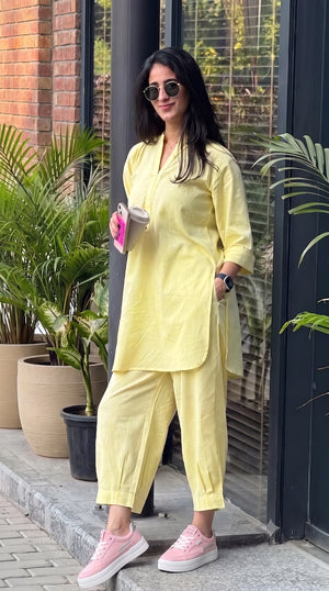 A woman wearing a light yellow two-piece kurta set with matching pants, sunglasses, and pink sneakers, standing outdoors in front of a glass door and potted plants.