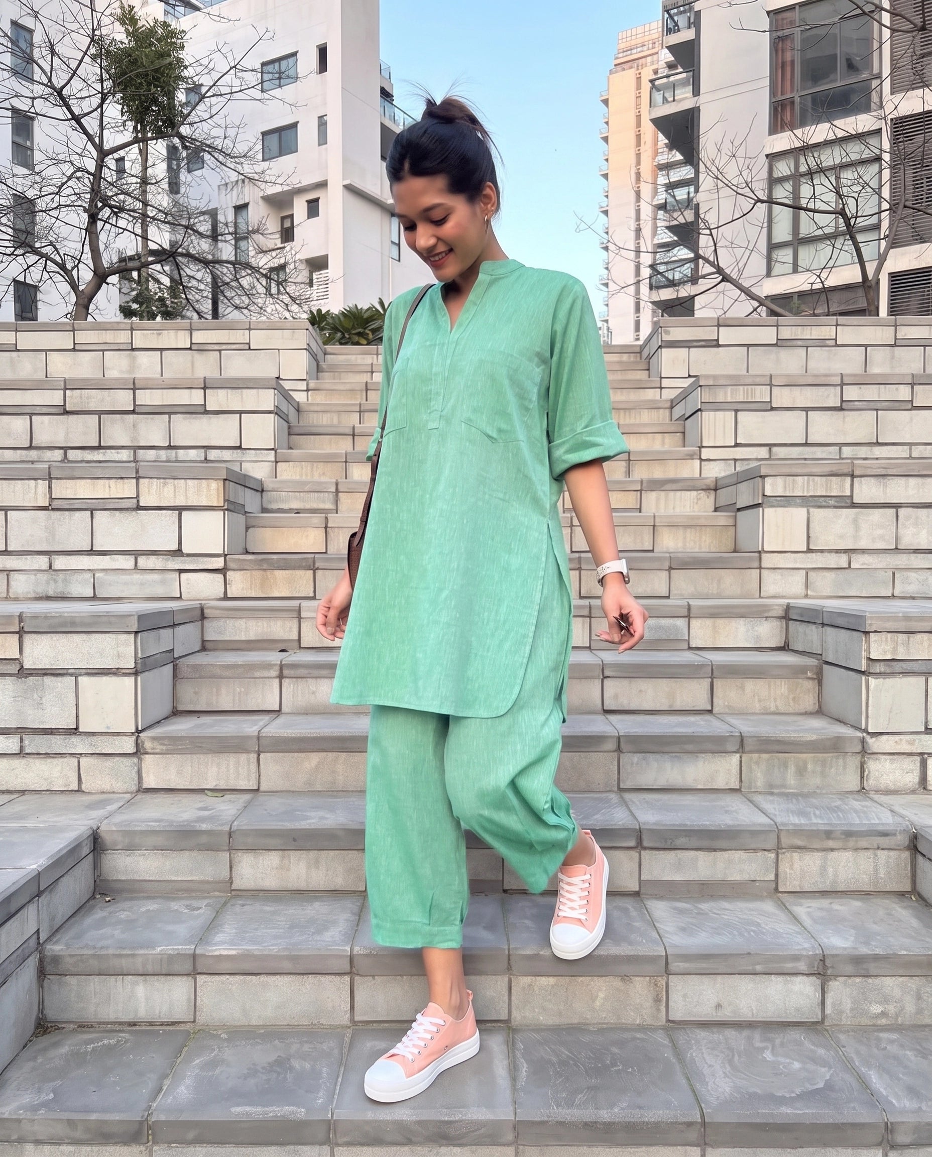 A woman wearing a light green two-piece linen set consisting of a short-sleeve tunic and wide-leg pants, walking down outdoor stone steps.