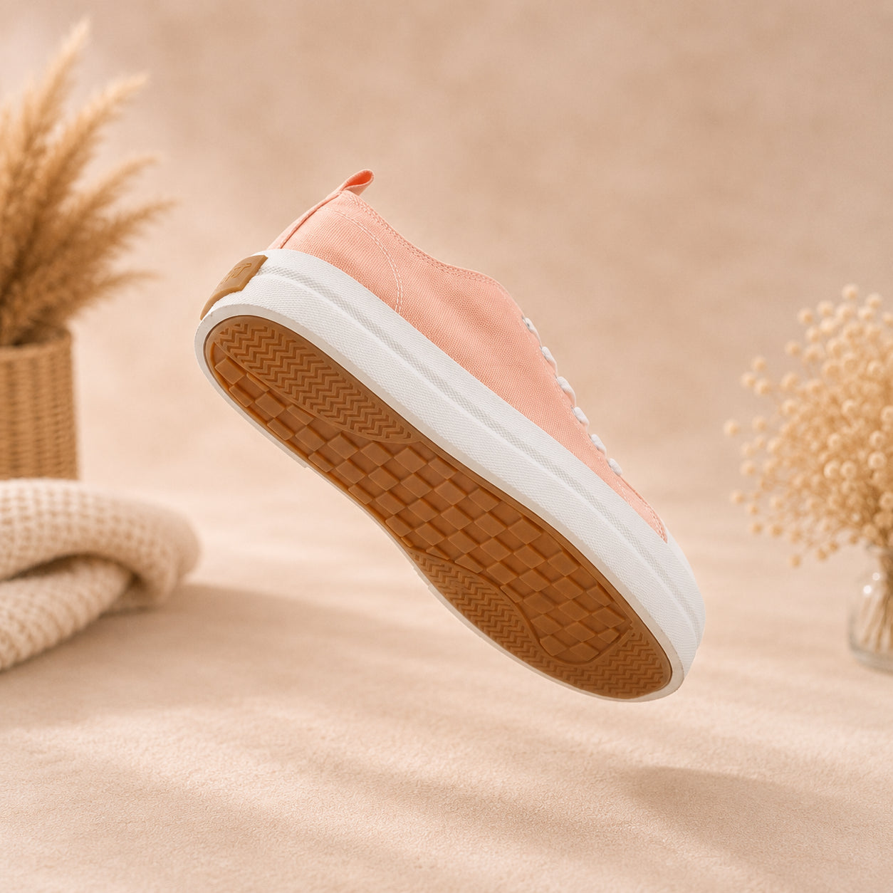 A peach-pink low-top canvas sneaker with a white midsole and a brown textured outsole, shown floating against a warm beige background with dried plants and a knitted textile.